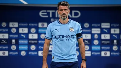A 35-year-old Portuguese assistant coach with a rugged build, wearing Manchester City's sky blue home shirt with navy...