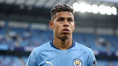 A young Brazilian winger in a sky-blue Manchester City training kit, waist-up, focused expression, blurred stadium...