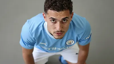 A 30-year-old Manchester City midfielder, wearing a blue jersey with a white collar, a white pair of shorts, and blue...