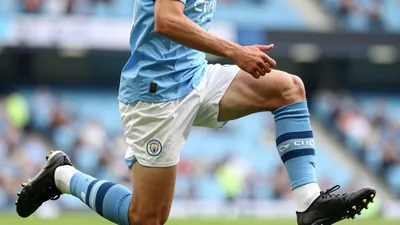 A 20-year-old Manchester City left-back, wearing a blue jersey and white shorts, with a black boot, is shown in a tight...
