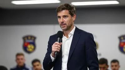 A 30-year-old football manager in a navy blue suit and white shirt, standing in a press conference room, speaking into a...