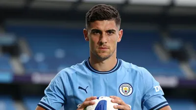 Mid-30s athletic midfielder, short dark hair, intense eyes, waist-up, wearing Manchester City sky-blue home shirt with...