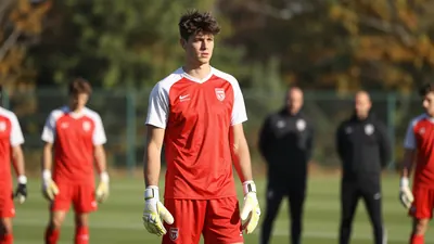 A 22-year-old, tall, athletic goalkeeper with a strong jawline and a messy, dark brown haircut, wearing a bright red...