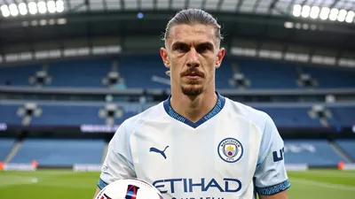 A photorealistic image of a Manchester City player in a white jersey with a blue collar, standing in front of a blurred...
