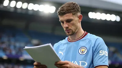A Manchester City midfielder in a sky blue jersey, standing in a stadium tunnel with a serious expression, looking at a...