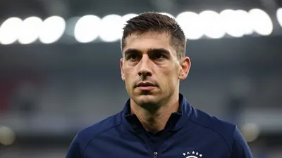 A mid-30s male City staff member in a navy blue training jacket, head-and-shoulders shot, background blurred stadium...